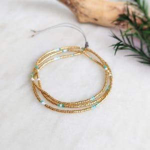 May include: Three gold beaded bracelets with turquoise and white accents are arranged in a circular pattern. The bracelets are strung on a gray cord, and the background is a marble surface.