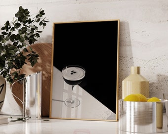 May include: A black and white photo of a cocktail in a glass with a black background. The photo is framed in a wooden frame and is sitting on a counter with a silver bowl of lemons and a silver pitcher.