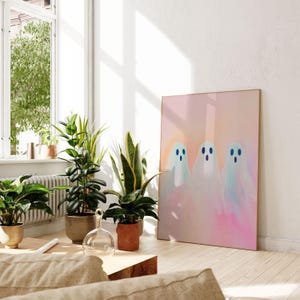 May include: A framed print of three white ghosts with blue eyes on a pink and orange gradient background. The print is hanging on a white wall in a room with plants.