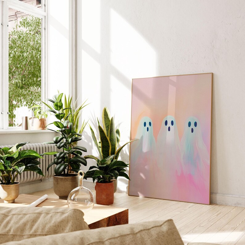 May include: A framed print of three white ghosts with blue eyes on a pink and orange gradient background. The print is hanging on a white wall in a room with plants.