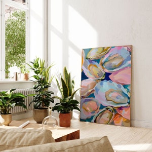 May include: Abstract painting of colorful oyster shells in shades of pink, blue, and yellow. The painting is framed and hanging on a white wall in a living room setting.