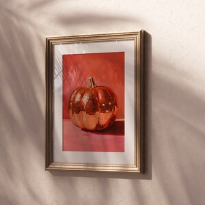 Pu&ograve; includere: Una stampa incorniciata di una zucca a forma di palla da discoteca dorata su sfondo rosa.