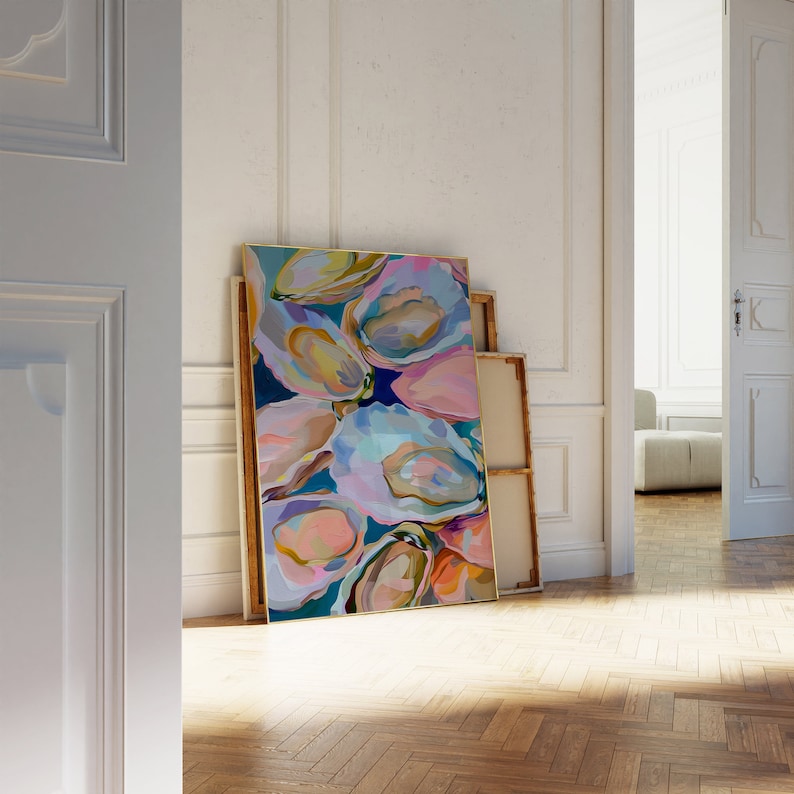 May include: Abstract painting of colorful oyster shells in shades of pink, blue, and yellow. The painting is framed in a gold frame and is leaning against a white wall.