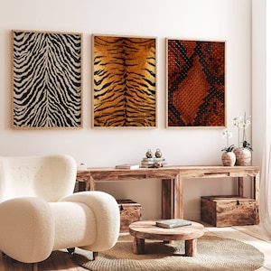 Op de afbeelding: Drie ingelijste prints van dierenprints. De prints zijn van een zebra, een tijger en een slang. De prints hangen aan een witte muur boven een houten tafel.