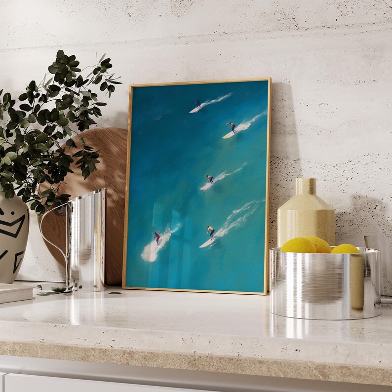 Puede incluir: Una pintura enmarcada de surfistas montando olas en un oc&eacute;ano turquesa. La pintura tiene un marco dorado y se exhibe en una encimera blanca.