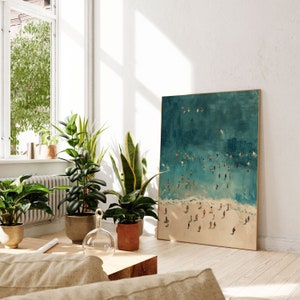 May include: A framed painting of a teal blue ocean with white waves and many small figures of people swimming and playing in the water. The painting is hanging on a white wall in a room with plants and a wooden table.