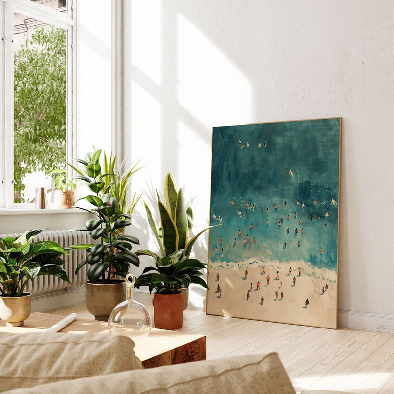 May include: A framed painting of a teal blue ocean with white waves and many small figures of people swimming and playing in the water. The painting is hanging on a white wall in a room with plants and a wooden table.