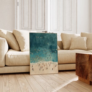 May include: A teal and white abstract painting of a beach scene with people swimming and sunbathing. The painting is framed in a gold frame.
