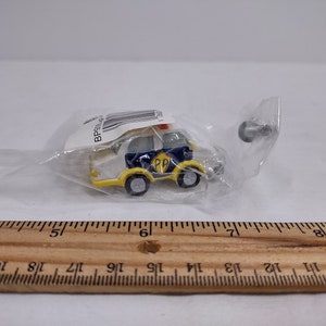 May include: A miniature police car toy, primarily yellow and navy blue, with a red light on top. The toy is packaged in a clear plastic bag. The car has the letters "PP" on the side. A small metal piece is also visible.