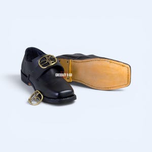 May include: A pair of black leather shoes with a square toe and a low heel. Each shoe features a prominent gold-toned metal buckle across the vamp. The text CRESSLEY & CO is printed in white between the shoes, which have light tan soles.
