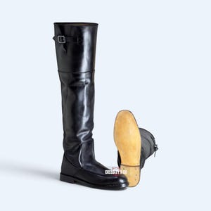 Op de afbeelding: Een paar zwarte kniehoge laarzen met een gladde afwerking en een zilverkleurige gesp aan de bovenkant. De ene laars staat rechtop terwijl de andere op zijn kant ligt, waardoor een lichtbruine zool zichtbaar is. De tekst CRESSLEY & CO is zichtbaar.