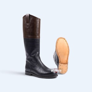 May include: A tall leather boot featuring a dark brown upper shaft and a black lower foot section. Beside it, a second boot is positioned to display its light tan, flat leather sole with visible stitching. The text CRESSLEY & CO is printed.