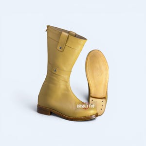 May include: A mid-calf yellow leather boot with a flat brown heel and a strap featuring a silver-tone metal stud. The boot has a smooth finish and a visible sole. The text CRESSLEY & CO is printed in white on a small rectangular background.