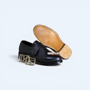 Puede incluir: Un zapato de cuero negro con una correa en la parte superior y dos hebillas de metal dorado a su lado. El zapato presenta una parte superior negra lisa y una suela de cuero color canela claro, con el nombre de la marca CRESSLEY & CO en blanco.