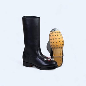 Puede incluir: Botas de montar de cuero negro con caña alta y tacón bajo. Una bota está de pie y la otra está en ángulo para mostrar la suela con tachuelas. Adecuadas para actividades ecuestres.