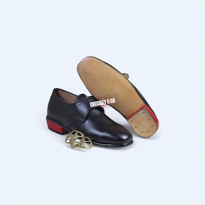 May include: Black leather shoes with red heels and soles, and gold-colored buckles. The sole of one shoe is visible, showing the wooden sole and the text "CRESSLEY & CO". The shoes are likely for dance or performance.