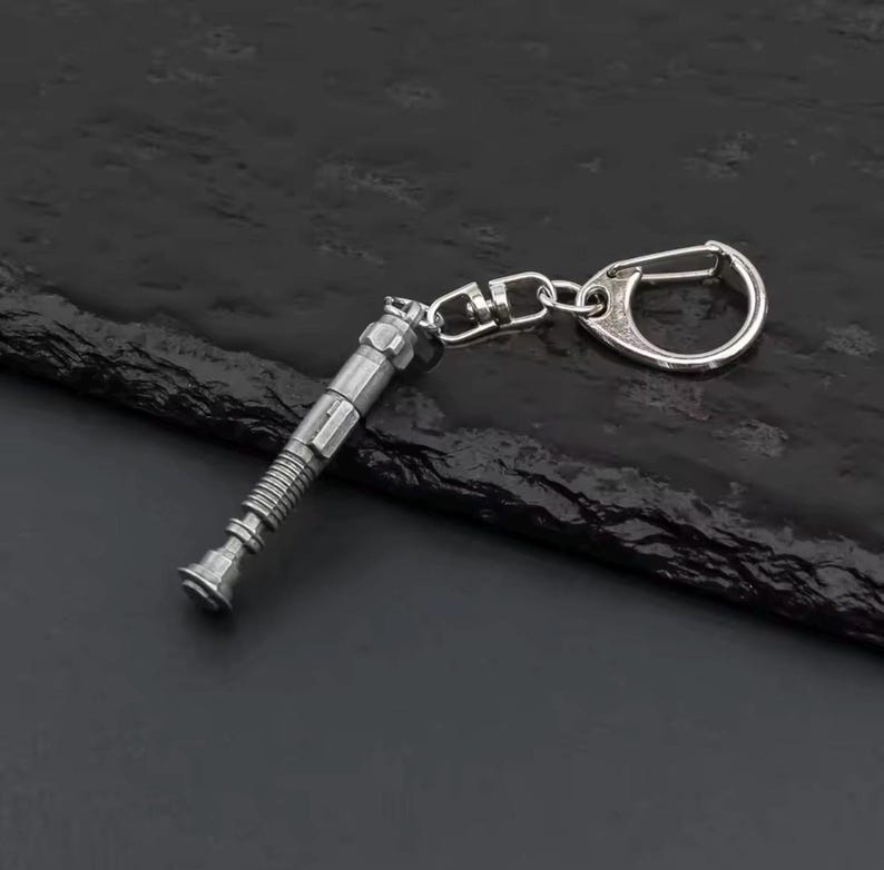 May include: A silver lightsaber keyring with a detailed hilt design and a keyring attachment. The keyring is made of metal and has a polished finish. The design is inspired by a popular science fiction franchise.