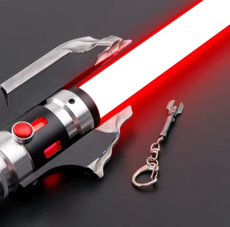 May include: A red and silver lightsaber with a bright red blade. The hilt is black and silver with a red button. A small silver wrench-shaped keyring hangs nearby. The lightsaber has a futuristic design.