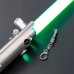 May include: A metallic silver lightsaber with a bright green blade. The hilt features intricate mechanical details, and a miniature keychain replica is attached. The lightsaber glows with a vivid green light.