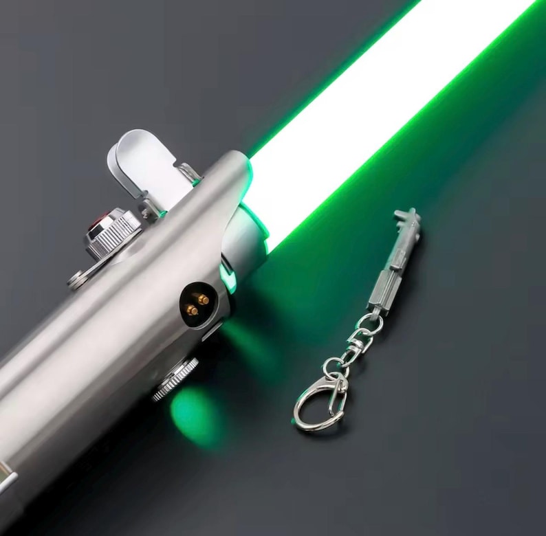 May include: A metallic silver lightsaber with a bright green blade. The hilt features intricate mechanical details, and a miniature keychain replica is attached. The lightsaber glows with a vivid green light.