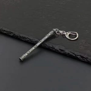 May include: A silver-coloured keychain with a detailed, cylindrical design. The keychain has a textured surface with various engraved patterns and a clasp for attaching keys. The design is reminiscent of a futuristic or fantasy-themed item.