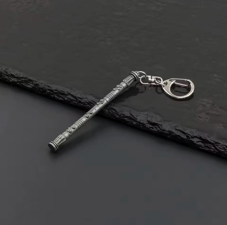 May include: A silver-coloured keychain with a detailed, cylindrical design. The keychain has a textured surface with various engraved patterns and a clasp for attaching keys. The design is reminiscent of a futuristic or fantasy-themed item.