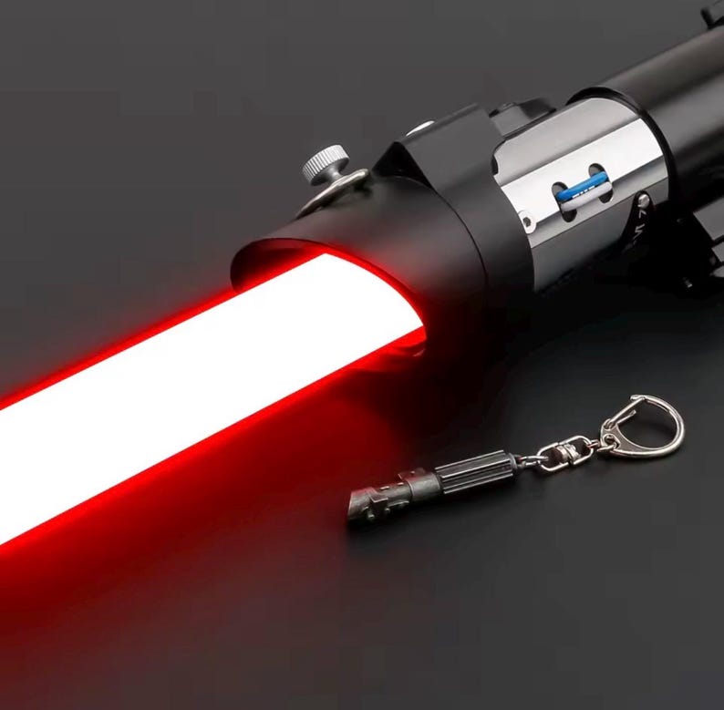 May include: A black lightsaber with a vibrant red blade, radiating a strong glow. A small keychain replica of the lightsaber is also visible. The lightsaber features silver accents and a detailed handle.