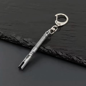 May include: A silver-coloured keychain featuring a lightsaber design. The keychain has a textured handle and a detailed hilt, with a split end. The keychain is attached to a silver ring and clasp.