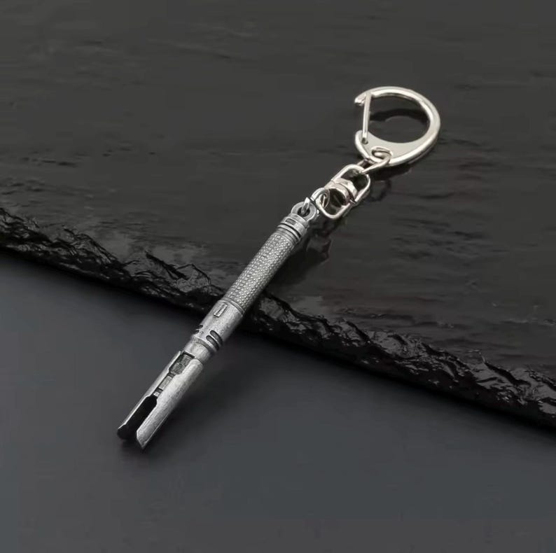 May include: A silver-coloured keychain featuring a lightsaber design. The keychain has a textured handle and a detailed hilt, with a split end. The keychain is attached to a silver ring and clasp.