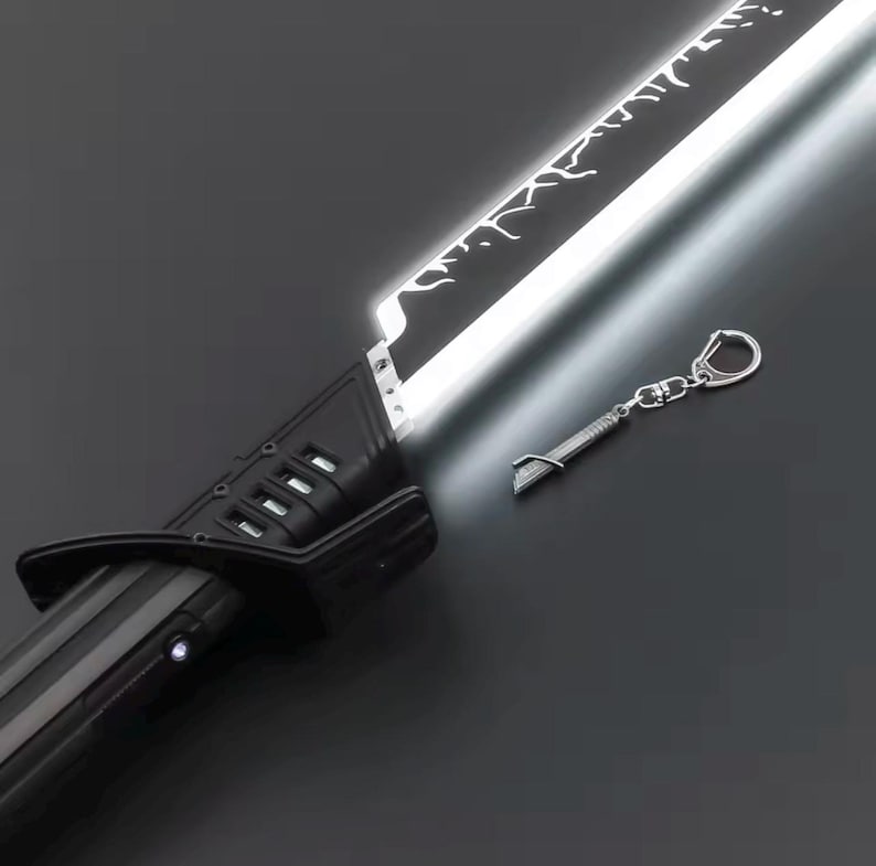 May include: A black and silver lightsaber with a bright white blade. The hilt has a sleek design with a few buttons. A small keychain replica of the lightsaber is also included, with a silver finish. The lightsaber is a prop or collectible.
