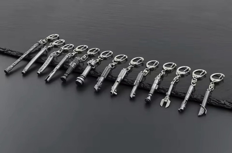 May include: A selection of silver lightsaber keyrings, each with a distinct design, displayed in a line. The keyrings are attached to silver key rings. The background is a dark, textured surface.