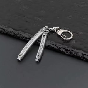 May include: Silver butterfly knife keychain. The knife is open, showing the blade and handle design. The handle has diamond-shaped engravings. The keychain has a silver clasp.