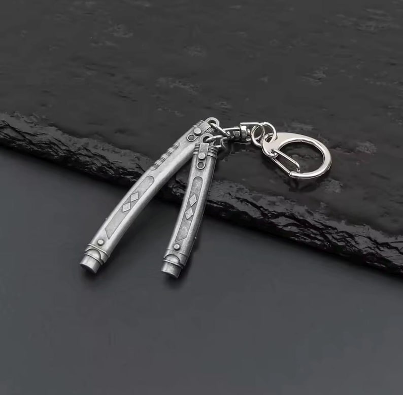 May include: Silver butterfly knife keychain. The knife is open, showing the blade and handle design. The handle has diamond-shaped engravings. The keychain has a silver clasp.