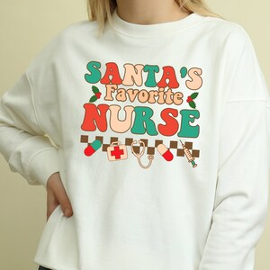 May include: White sweatshirt with a retro-style graphic that says "Santa's Favorite Nurse" in red, orange, green, and blue. The graphic also includes a red cross, a stethoscope, a pill, and a syringe.