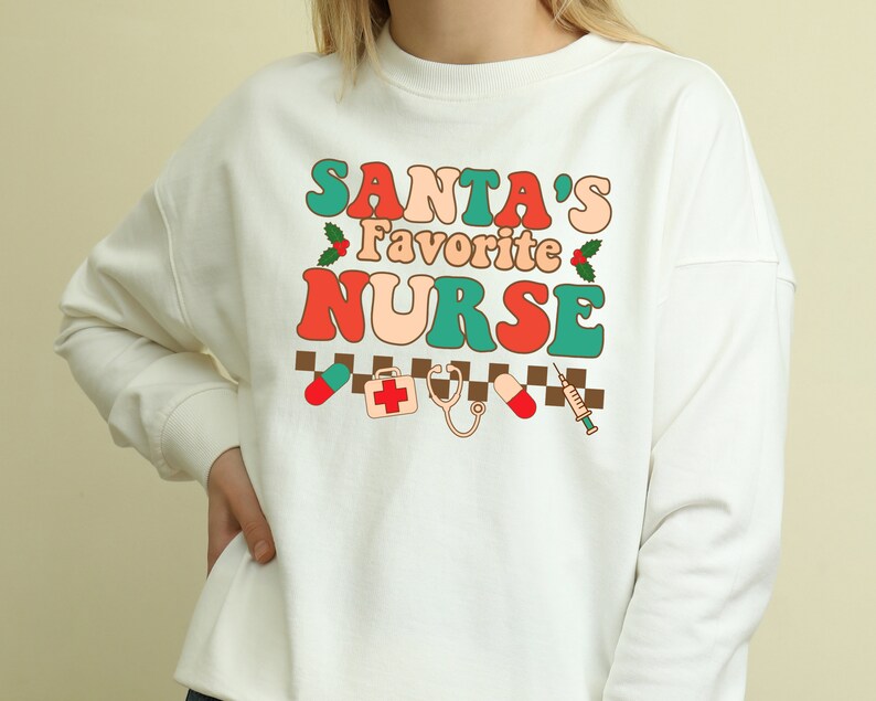 May include: White sweatshirt with a retro-style graphic that says "Santa's Favorite Nurse" in red, orange, green, and blue. The graphic also includes a red cross, a stethoscope, a pill, and a syringe.