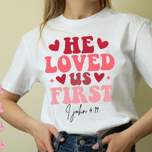 Puede incluir: Camiseta blanca con un gráfico rosa y rojo que dice "He Loved Us First" con corazones y una pequeña estrella. El texto "1 Juan 4:19" está escrito en cursiva debajo del gráfico.