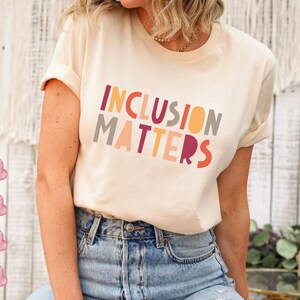 May include: A woman wearing a cream colored t-shirt with the text "INCLUSION MATTERS" in colorful block letters. The text is in a rainbow color scheme.