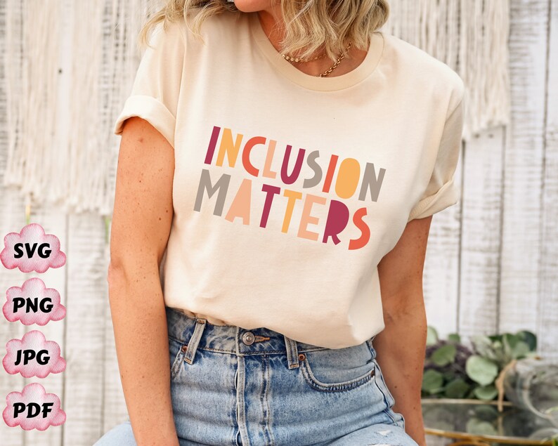 May include: A woman wearing a cream colored t-shirt with the text "INCLUSION MATTERS" in colorful block letters. The text is in a rainbow color scheme.