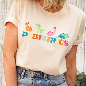 May include: A white t-shirt with the word "PEDIATRICS" spelled out in colorful letters. Each letter has a small cartoon dinosaur on it. The dinosaurs are green, orange, pink, and blue.