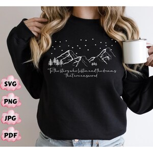 Op de afbeelding: Zwart sweatshirt met een wit grafisch ontwerp van bergen, sterren en bomen. De tekst "To the stars who listen and the dreams that are answered" staat op het sweatshirt gedrukt.