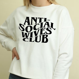 May include: White long-sleeve sweatshirt with black text that reads "ANTI-SOCIAL WIVES CLUB" in a retro font.
