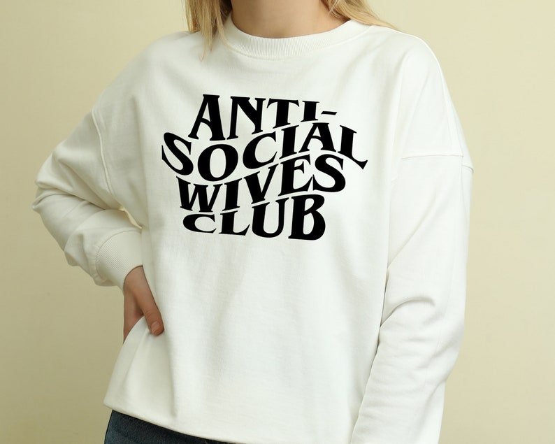 May include: White long-sleeve sweatshirt with black text that reads "ANTI-SOCIAL WIVES CLUB" in a retro font.