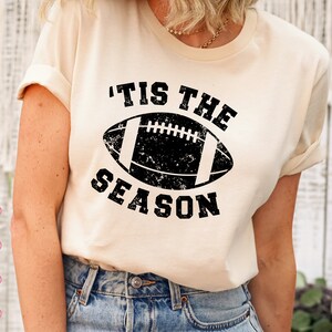 May include: A white t-shirt with a black distressed graphic of a football and the text 'Tis the Season'.