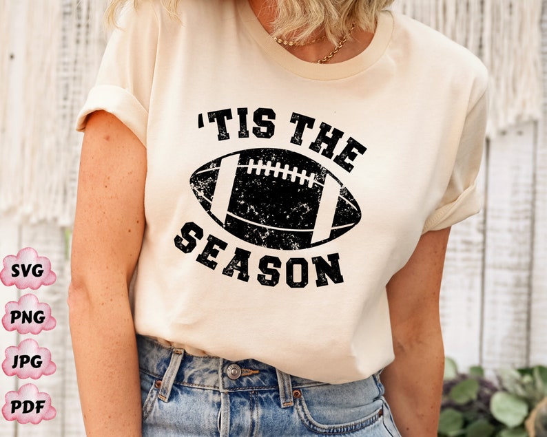 May include: A white t-shirt with a black distressed graphic of a football and the text 'Tis the Season'.