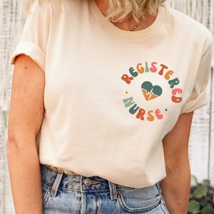 Peut inclure: Un t-shirt blanc avec le texte "REGISTERED NURSE" dans une police r&eacute;tro color&eacute;e. Un c&oelig;ur avec un graphique de battement de c&oelig;ur est au centre du texte.