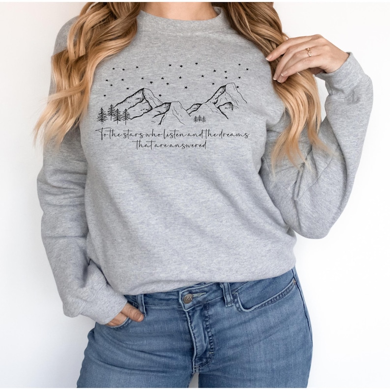 Op de afbeelding: Grijs sweatshirt met een zwarte bergketen en sterren graphic. De tekst "To the stars who listen and the dreams that are answered" is onder de graphic gedrukt.