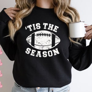 May include: Black sweatshirt with a white distressed graphic of a football and the text 'Tis the Season'.