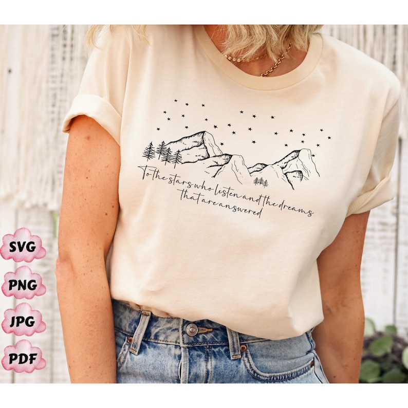 Op de afbeelding: Een beige T-shirt met een zwart grafisch ontwerp van bergen, sterren en bomen. De tekst "To the stars who listen and the dreams that are answered" staat op het shirt gedrukt.