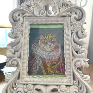 May include: Ornate, white picture frame with a portrait of a cat wearing a crown and regal attire. The frame has a detailed, scrolling design with floral accents. The cat portrait features a green background and gold embellishments.