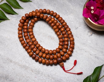 May include: A brown wooden mala bead necklace with a red tassel. The mala beads are strung on a cord and have a dark brown wooden bead at the end.